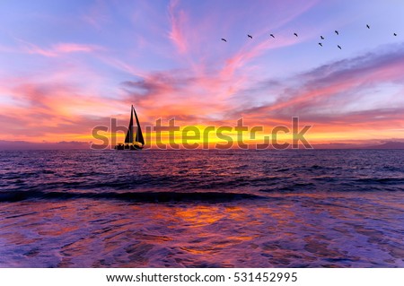 Ocean sunset sailboat silhouette is sailboat sailing along the ocean water with a colorful vivid sunset sky and silhouettes of birds flying in the background..