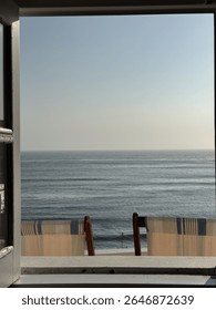 Ocean view through an open window with striped deck chairs on the balcony, overlooking the Atlantic coast in Algarve, Portugal on a sunny summer day.