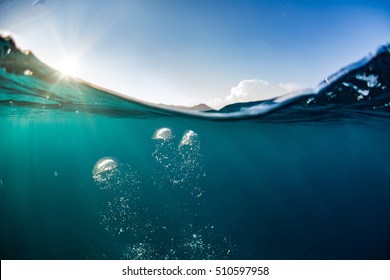 Ocean Water Line Splits Sky And Underwaer Part. Air Bubbles In Deep Blue. Bright Sun Light And Blue Clear Sky