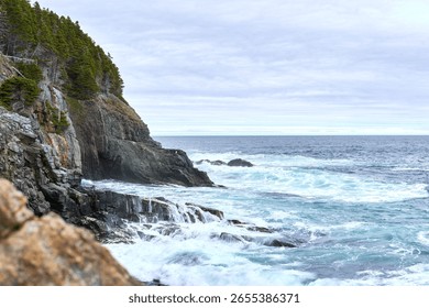 The ocean waves create foam while touching the rocks of a cliff under the cloudy blue sky