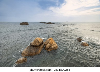 Ocean Waves Gently Lap Against Large Rocks Under a Softly Clouded Sky at a Coastal Destination. The coast of the port city of Galle on the south-west coast of Sri Lanka.