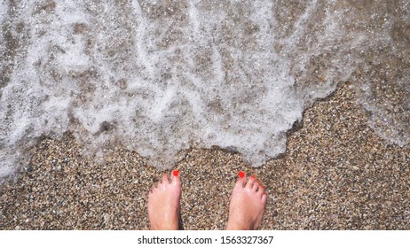 Ocean Waves Washing Over Slim Female Feet. Woman Standing On Seashore With Pebble And Enjoying Tides Of Clear Sea Water. Young Girl Enjoying Summer Vacation At Seaside. Trip Concept. Slow Mo POV