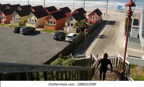 Oceanside, California USA - 20 Feb 2020: Surfer man with surfboard going surfing ocean waves. Colorful waterfront cottages and lifeguard tower, watchtower. Multicolor bungalow huts by sea. Palm trees.