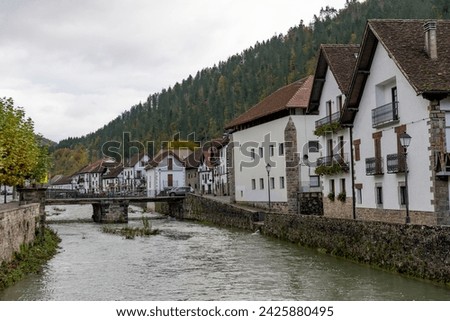 Ochagavia townscape on cloudy autumn  day overlooking typical residential buildings with steep brown tiled roofs along stone embankment and bridge across Anduna river on sunny day, Navarra, Spain