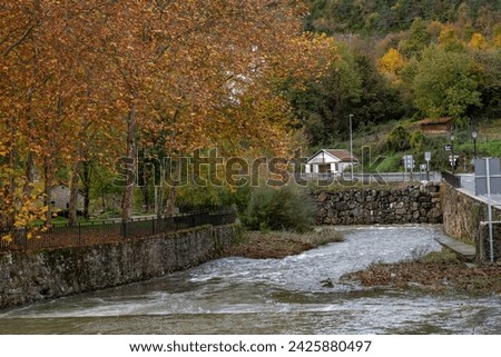 Ochagavia townscape on cloudy autumn  day overlooking typical residential buildings with steep brown tiled roofs along stone embankment and bridge across Anduna river on sunny day, Navarra, Spain