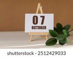 October 7th. Day 7 of month, Calendar date. Green branch, easel with the date and month on desktop. Close-up, brown background. Autumn month, day of year concept