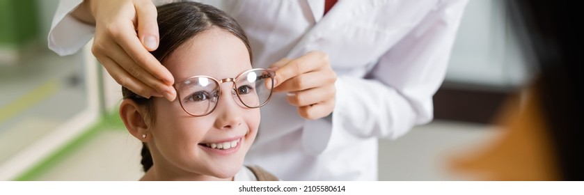 Oculist Trying Glasses On Smiling Girl Near Blurred Mother In Optics Store, Banner