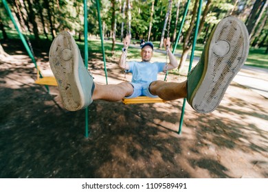 Odd bizarre eccentric people concept. Adult stylish man in glasses with crazy funny emotional  face riding on swing in city park on playground for children in summer.  Happy person enjoying childhood