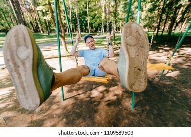 Odd bizarre eccentric people concept. Adult stylish man in glasses with crazy funny emotional  face riding on swing in city park on playground for children in summer.  Happy person enjoying childhood
