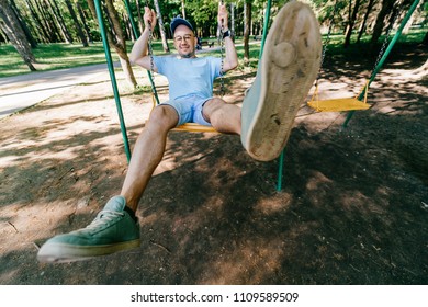 Odd bizarre eccentric people concept. Adult stylish man in glasses with crazy funny emotional  face riding on swing in city park on playground for children in summer.  Happy person enjoying childhood