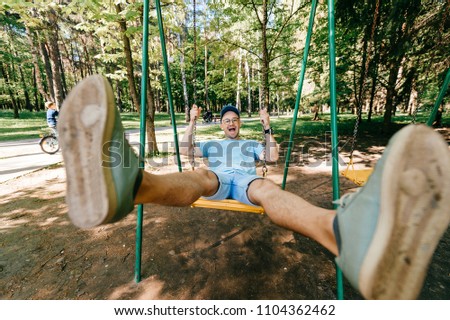 Odd bizarre eccentric people concept. Adult stylish man in glasses with crazy funny emotional  face riding on swing in city park on playground for children in summer.  Happy person enjoying childhood