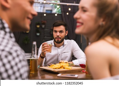 Odd Friend. Calm Young Man Thoughtfully Drinking His Alcohol And Looking At Two Friends Flirting