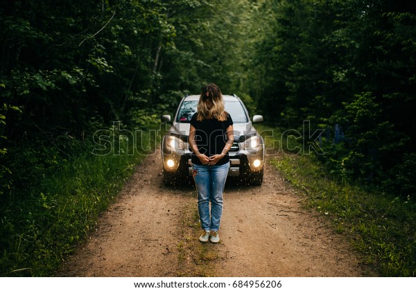 Odd, strange, unrecognizable and unknown person woman\
with long lush hair on face standing alone in front of big car on\
lost nameless road in gloomy dark rainy forest.  Horror movie\
concept. Scary. 