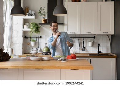 Ode To Passion For Food. Happy Millennial Husband Standing By Table At Modern Kitchen Cooking Romantic Dinner To Beloved Wife. Overjoyed Young Man Singing In Imaginary Microphone Made Of Egg Beater