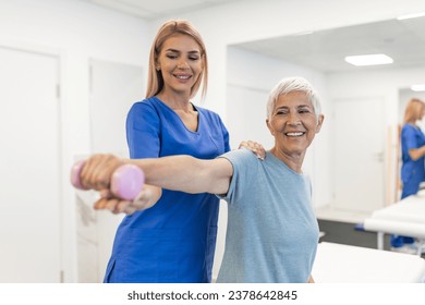 Oder aged woman doing physiotherapy with support from physiotherapists . Senior elderly female sitting in clinic using dumbbells workout exercise for patient with caregiver in nursing care.