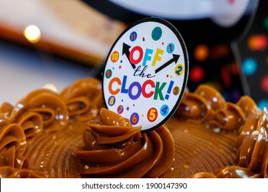 Off The Clock Cake Topper Decoration That Looks Like A Timeclock With Hour And Minute Hands On Top Of Caramel Chocolate Whipped Icing Yellow Rich Cheesecake At Retirement Party With Bokeh Background.