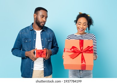 Offended African American Man With Small Gift Box Looking At Amazed Wife With Big Present On Blue