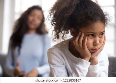 Offended African American preschooler girl looking in distance, holding head in hands, talking mother on background, family conflict, child punishment, complicated relationships with parent, close up
