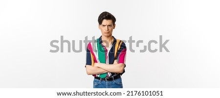 Offended and angry young gay man sulking, cross arms chest and looking judgemental at camera, standing over white background