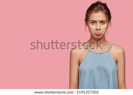 Offended Caucasian girl with insulted expression, scowls face, puses lips in discontent, has appealing look, dressed in casual clothes, poses over pink background, copy space for your advertisement