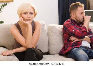 Offended Couple Man And Woman Do Not Speak To Each Other While Sitting On Sofa At Home. Disappointed And Sad Blond Lady Looking At Camera.