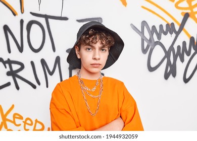 Offended displeased male teenager keeps hands crossed looks unhappily wears black hat orange t shirt metal chains around neck poses against graffiti wall. Modern youth and urban style concept