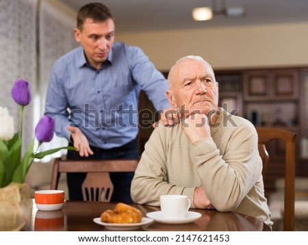 Offended elderly man sitting at table with disgruntled adult son standing behind and reprimanding him