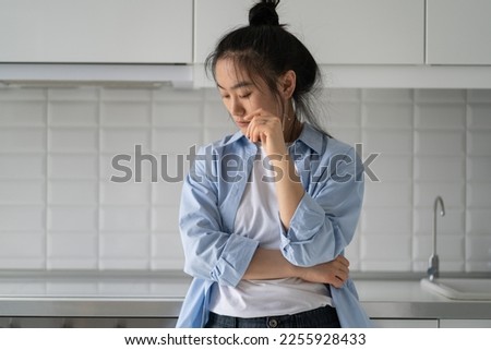 Offended embarrassed Asian woman looking down and feeling sad after breaking up with boyfriend. Discouraged depressed Korean girl standing in kitchen after stressful conversation with husband