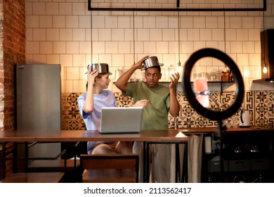 Offended Girl Looking On Black Guy During Making Video In Prank Or Challenge On Smartphone For Social Networks. Multiracial Blogger Couple Wearing Saucepan On Heads At Table On Home Kitchen