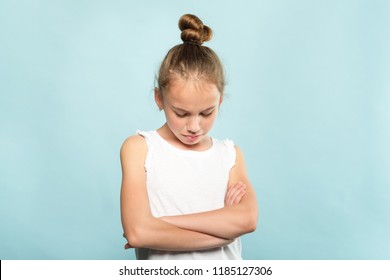 offended grumpy hurt child with fold hands looking down. young cute huffy girl portrait on blue background.