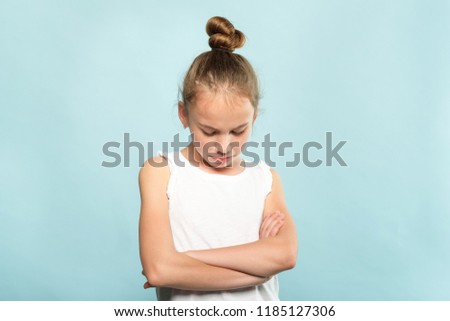 offended grumpy hurt child with fold hands looking down. young cute huffy girl portrait on blue background.