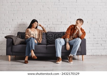 offended interracial couple looking at each other on couch in living room, relationship difficulties. Translation of tattoo: be faithful to the one who is faithful to you
