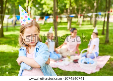 Offended littlee child hugging herself and scowling on background of other people enjoying picnic in park on summer day