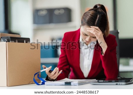 In the office, an Asian woman carefully signs her resignation letter, feeling stressed. She packs her belongings in cardboard boxes, preparing to leave the workplace. Asian people.