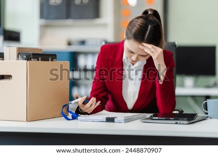 In the office, an Asian woman carefully signs her resignation letter, feeling stressed. She packs her belongings in cardboard boxes, preparing to leave the workplace. Asian people.