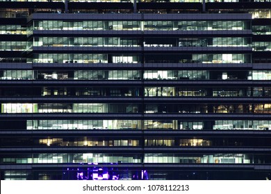 Office Building At Night. Late Night At Work. Glass Curtain Wall Office Building. Blue Light Shining On Building Elevation Facade.