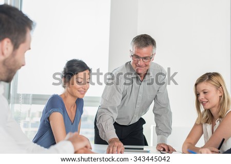 Office business meeting. The team is sitting at a table in a luminous white open space The men are wearing suits and shirts. Everybody is watching the senior grey haired boss blueprint