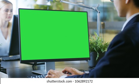 In The Office Businessman Works At His Desk On A Personal Computer With Mock-up Green Screen. His Colleague Sets Opposite To Him.