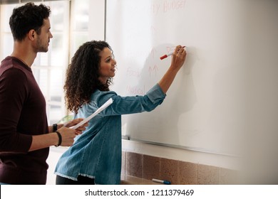 Office colleagues discussing monthly budget and plans on a whiteboard. Businesswoman writing on the board while her colleague holds a paper in hand.