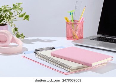 Office concept photo from front view, over white background. In the middle of white tabletop contains some notebook and clipboard, next to a laptop, pen pot, and a pink headphone. Space for designing - Powered by Shutterstock