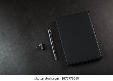 Office Desk With Black Notepad And Pen On Black Background.