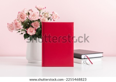 Office desk, red book cover mockup, vase with pink flowersroses on pink wall background. Front view. Place for text, copy space, mockup