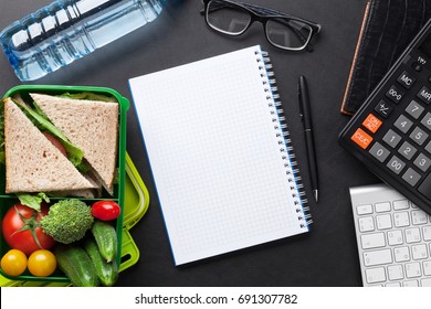 Office Desk With Supplies And Lunch Box With Vegetables And Sandwich. Top View With Notepad For Your Text