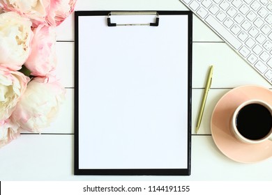 Office desk table with cup of coffee, pink peony flowers, golden pen, blank paper and clipboard. White wooden background. Coffee break, ideas, to do list, plan or note writing concept. Top view.