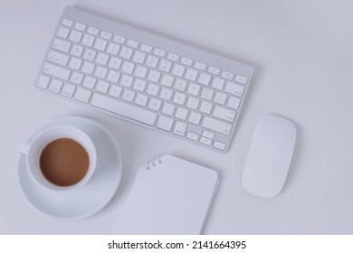 Office desk table with keyboard, blank note, mouse trackpad and coffee cup. All in white, clean and minimalist style. Top view with copy space