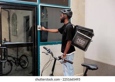 Office food delivery service courier pressing company building doorbell, waiting for customer outdoors. African american man delivering restaurant takeaway lunch, standing in front of door - Powered by Shutterstock