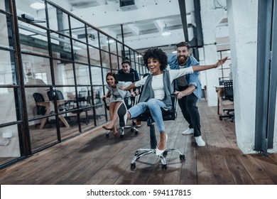 Bürospass. Vier junge, fröhliche Geschäftsleute in intelligenter, ungezwungener Kleidung, die Spaß beim Rennen auf Bürostühlen und Lächeln haben