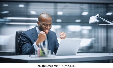 Office: Happy Successful Black Businessman Sitting At Desk Using Laptop Computer, Celebrate Success. Entrepreneur In Suit Working With Stock Market App Smiles, Happy Victory. Motion Blur Background.