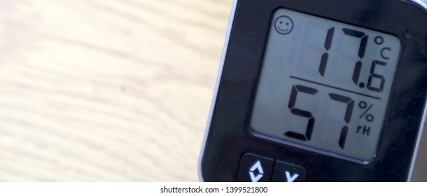 Office Or Home Temperature Gauge Showing Temperature And Humidity, Black In Colour On A Light Coloured Surface Desk, Displaying 17.6 Degrees Celsius And 57% Humidity With A Smile Icon In Top Left.