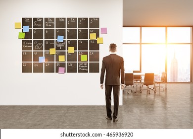 Office interior. Businessman standing in room looking at large calendar on wall. Concept of good scheduling. 3d rendering. Toned image